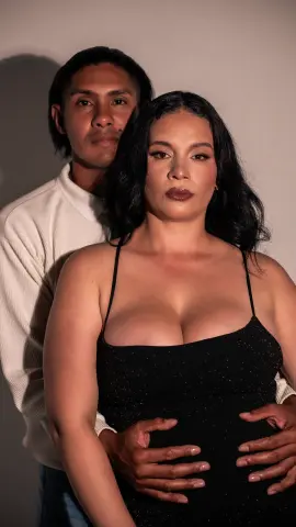 Romantic couple in elegant evening wear posing against a neutral studio background with moody lighting.