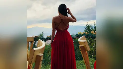 Woman in a long red evening gown standing on a balcony overlooking a scenic mountain landscape at dusk.