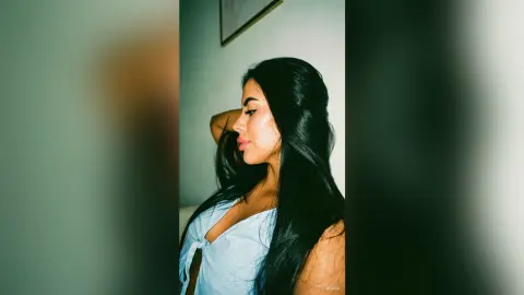Beautiful woman with long dark hair wearing a light blue tie-front top in a soft, moody indoor setting.