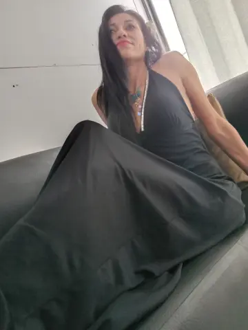 Elegant woman in a long black halter dress relaxing on a sofa in a bright indoor setting.