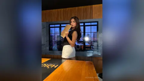 VictoriaLorent poses in a black sleeveless top and white mini skirt, holding a tan folder against her chest in a room with a city view window.