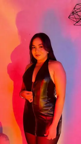Woman in a black leather halter dress posing under warm pink and orange studio lighting.