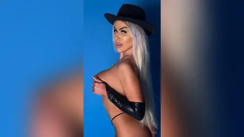 Glamorous blonde woman in a black cowboy hat and leather gloves posing against a blue studio background.