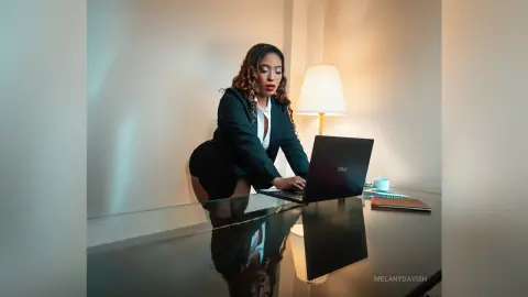 Professional woman in a black blazer working on a laptop at a glass desk in a warm, modern office setting.