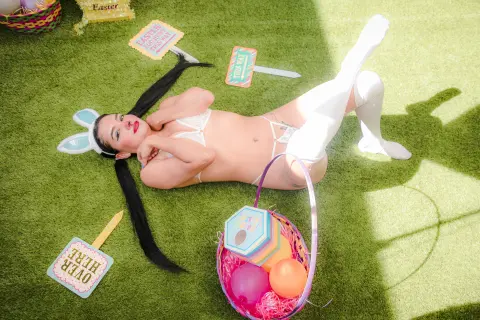 Woman in white bunny ears and lingerie lying on green grass with colorful Easter egg baskets.