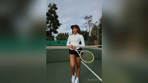 Athletic woman in white long-sleeve tennis shirt and black skirt holding a racket on an outdoor tennis court.