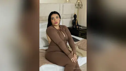 Curvy woman in a brown long-sleeve bodysuit relaxing on a bed in a cozy bedroom setting.