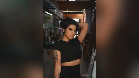 Young woman with short dark hair in a black crop top posing in a moody, dimly lit urban setting.