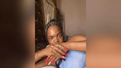 Young woman with braided hair and red nails relaxing in a cozy bedroom setting.