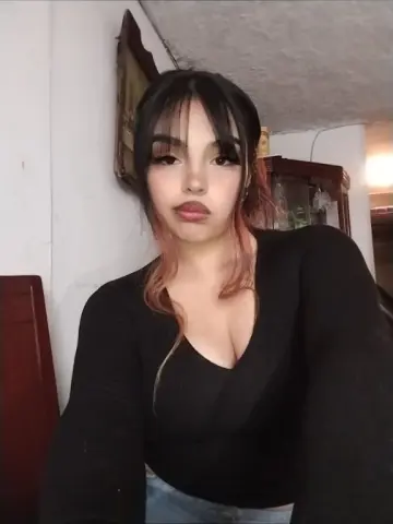 Young woman with long dark hair and bangs wearing a black long-sleeve top in a cozy indoor setting.