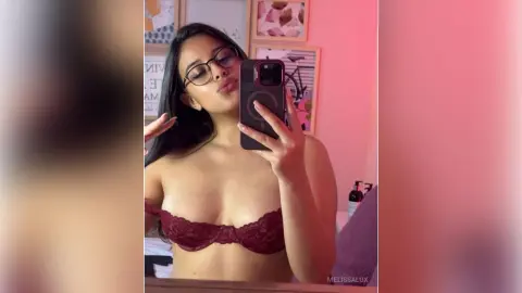 Young woman with glasses in burgundy lace lingerie taking a selfie in a cozy, pink-lit bedroom.