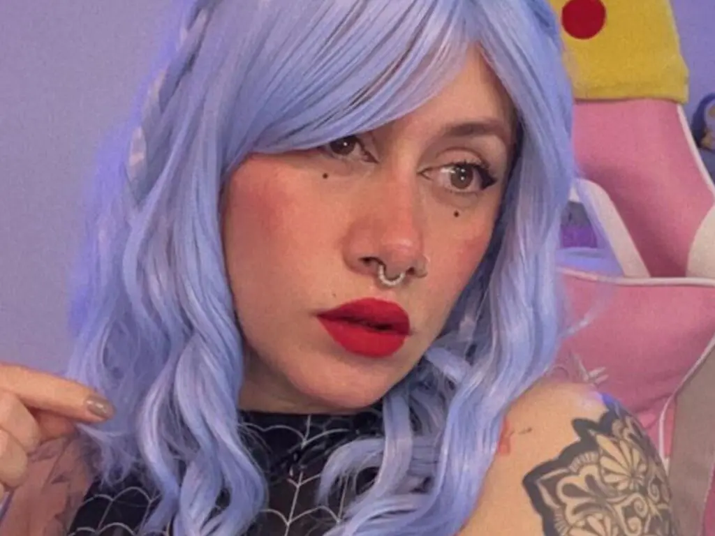 Woman with wavy blue hair, red lipstick, and nose ring wearing a black mesh outfit in a pastel pink room.