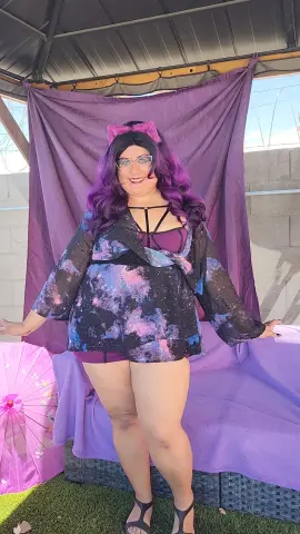 Smiling woman with purple hair in a galaxy print blouse and pink bow posing against a purple backdrop.
