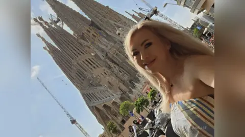 Blonde woman smiling in front of the Sagrada Familia cathedral in Barcelona under a bright blue sky.