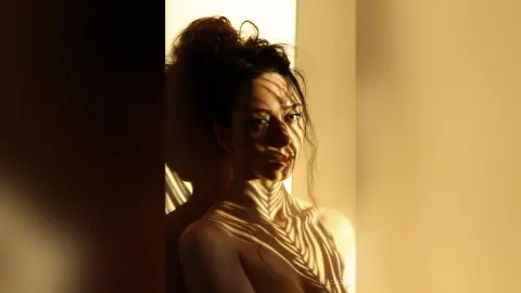 Moody portrait of a woman with dramatic sunlight and leaf shadows cast across her face and skin.