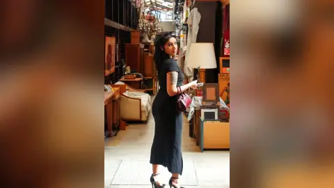 Stylish woman in a black midi dress standing in a vintage antique shop filled with retro decor.