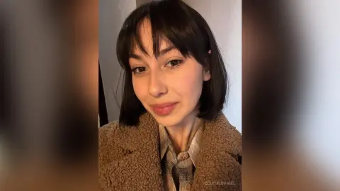 Young woman with dark bangs wearing a cozy brown teddy coat and plaid shirt in a soft indoor setting.