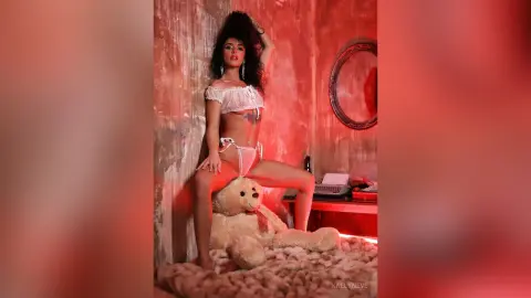 Glamorous woman in white lace lingerie sitting on a teddy bear in a moody, red-lit room.