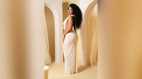 Elegant woman in a white backless evening gown posing in a minimalist cream arched architectural setting.