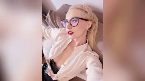 Glamorous blonde woman in glasses and a beige blouse relaxing on a sofa with a chic, confident mood.