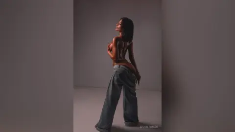 Slim woman in baggy denim jeans posing in a studio with moody, dramatic lighting.