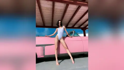 Fashionable woman in a white bodysuit leaning against a vintage pink car under a wooden pergola on a sunny day.