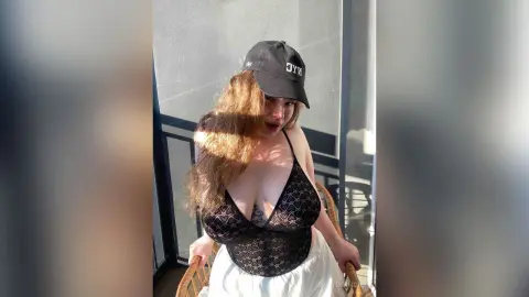 Young woman in a black lace bodysuit and baseball cap sitting on a sunny balcony.