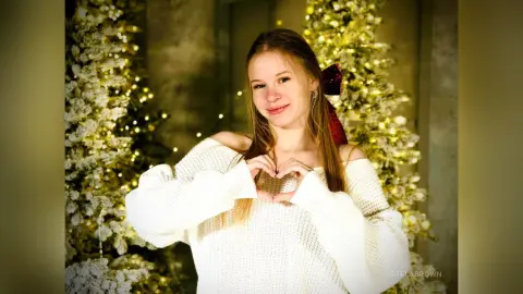 StelaBrown smiles while wearing an off-the-shoulder white sweater, posing in front of a lit Christmas tree.
