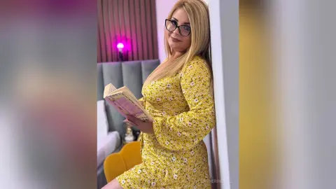 Blonde woman in a yellow floral dress reading a book in a cozy, brightly lit bedroom setting.
