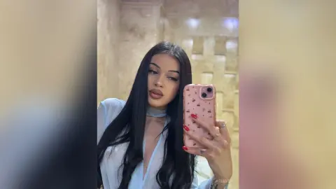 AriaVex takes a mirror selfie wearing a white open-front top with long black hair.