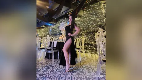 MeganneAllen poses in a black slit dress and strappy heels, standing in a room with clear chairs and warm fairy lights.