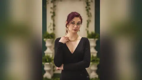 Elegant woman with red hair in a black V-neck dress and gold necklace posing in a bright garden setting.