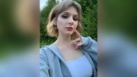 AlitaPatti poses outdoors with her hand near her chin, wearing a white tank top and a grey zip-up hoodie.