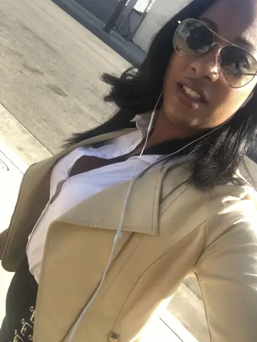 LondonSquirts wears sunglasses and a white shirt under a tan jacket while standing outdoors.