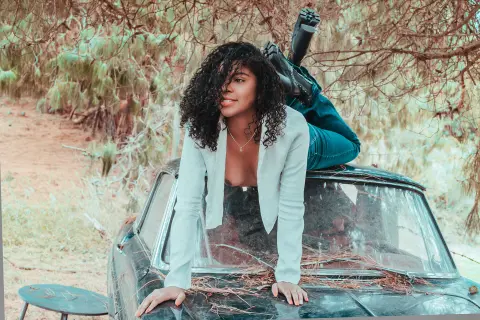 Curvy woman in a white blouse leaning on a vintage car in a dreamy, sunlit forest setting.