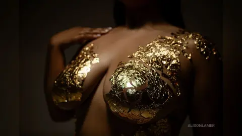 Woman with shimmering gold leaf body art on skin in a moody, dramatic studio setting.