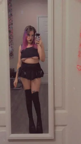 Young woman with pink hair in a black crop top and mini skirt posing for a mirror selfie in a bedroom.