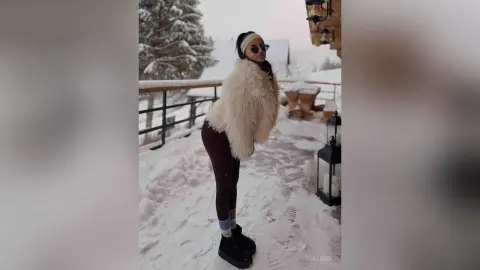 Stylish woman in a white faux fur coat and sunglasses posing on a snowy winter balcony at a mountain resort.
