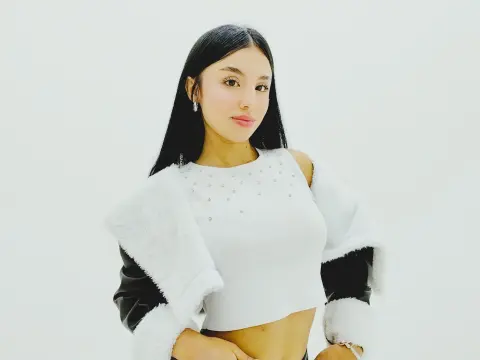 Ema_Velvet poses in a white cropped top with white faux fur trim and long black hair against a plain white background.