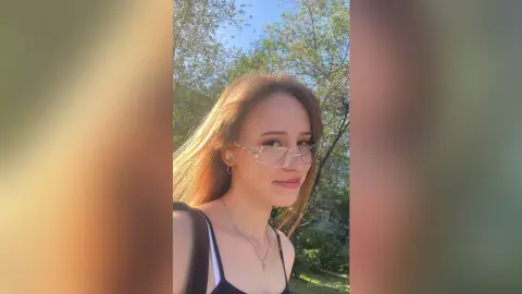 Smiling young woman with glasses and long hair posing outdoors in soft natural sunlight.
