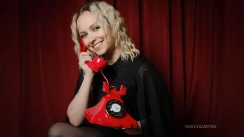 Smiling blonde woman in a black dress holding a vintage red rotary telephone against a red curtain backdrop.