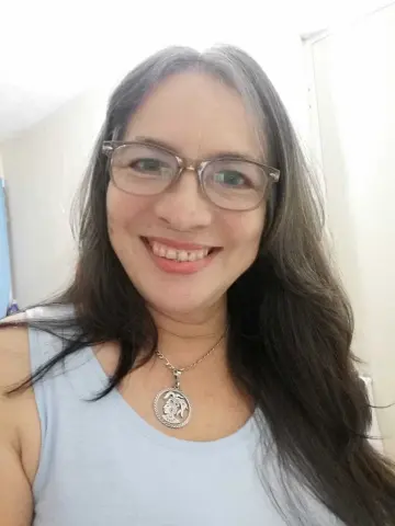 Smiling woman with long dark hair wearing glasses and a light blue tank top and silver medallion necklace.