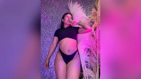 Young woman in black crop top and briefs posing against a sparkly backdrop with pink lighting and pampas grass.