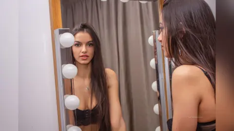 Young woman with long dark hair in a black top looking into a lighted vanity mirror in a dressing room.