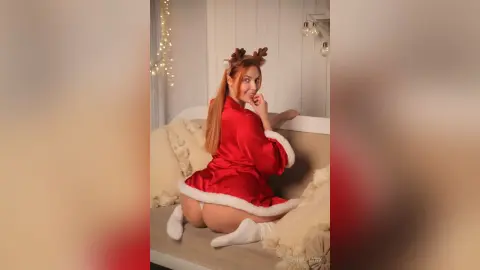 Smiling woman in a red satin Santa outfit sitting on a cozy white sofa in a festive holiday setting.