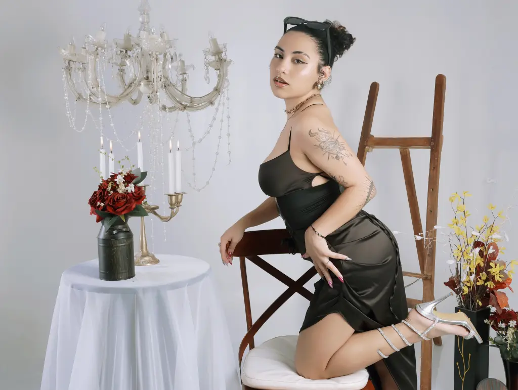 BrianaKnox — Elegant woman in a black satin dress posing by a vintage table and chandelier in a chic studio setting.