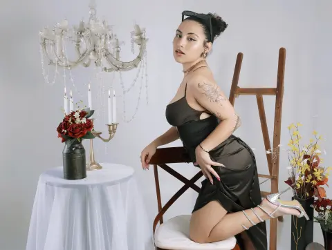 BrianaKnox — Elegant woman in a black satin dress posing by a vintage table and chandelier in a chic studio setting.
