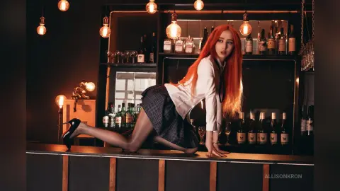 AllisonKron poses in a white blouse and plaid skirt, leaning back against a bar counter in black sheer tights and heels.