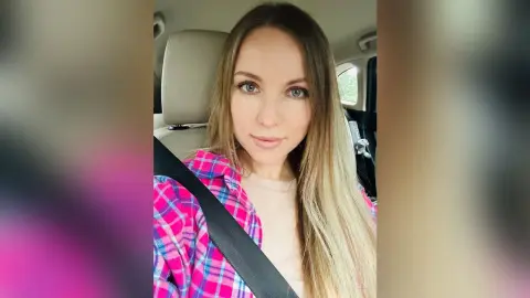 Smiling young woman with long blonde hair wearing a pink plaid shirt sitting in a car seat.