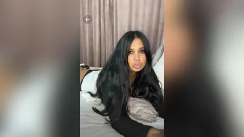 Young woman with long dark hair wearing a white top relaxing on a bed in a cozy bedroom setting.